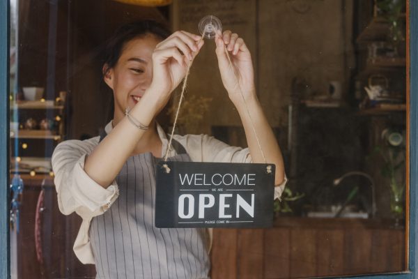 Young Asia manager girl changing a sign from closed to open sign on door cafe looking outside waiting for clients after lockdown. Owner small business, food and drink, business reopen again concept.
